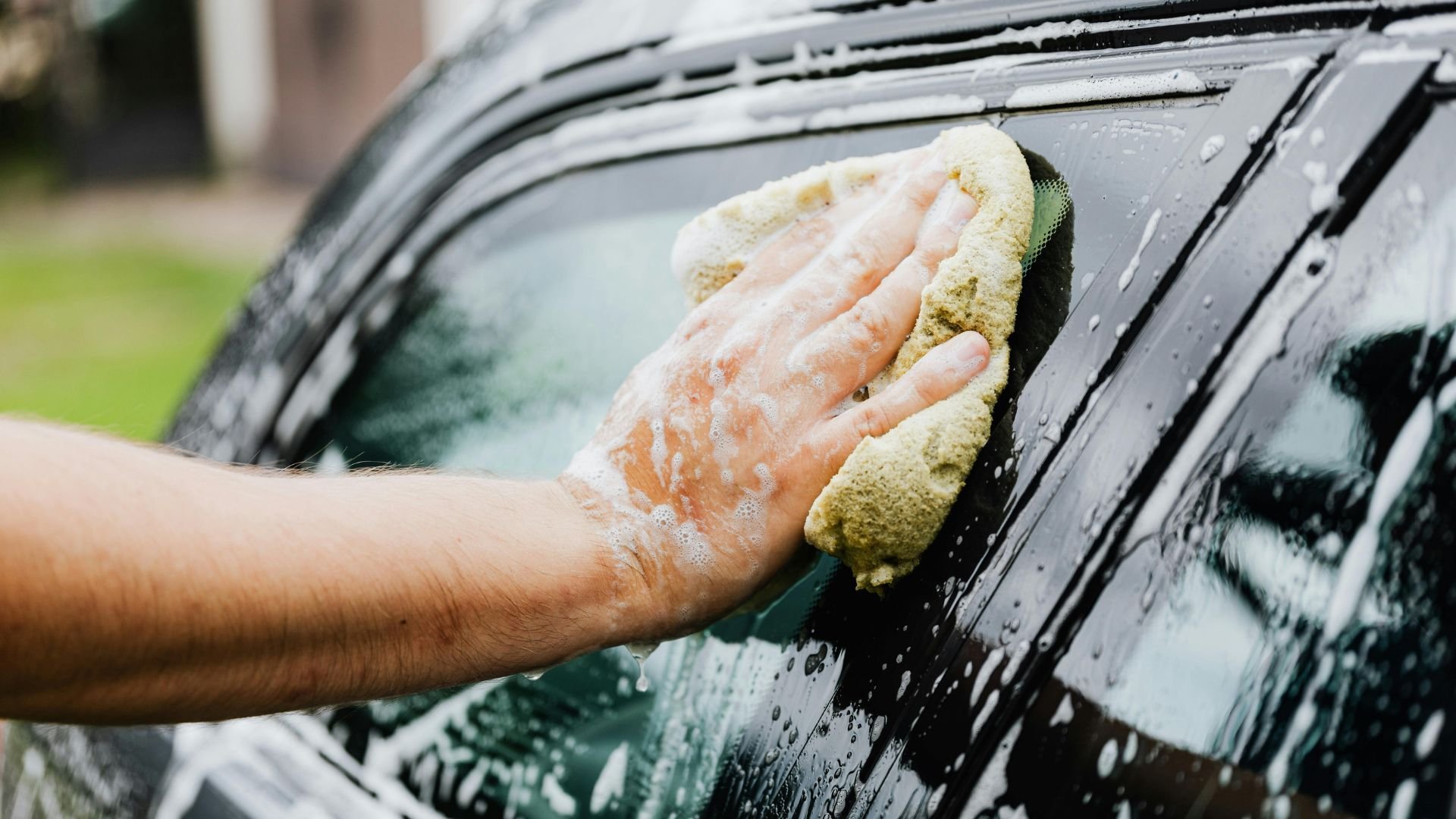 Hand washing car window with soapy sponge, water droplets visible
