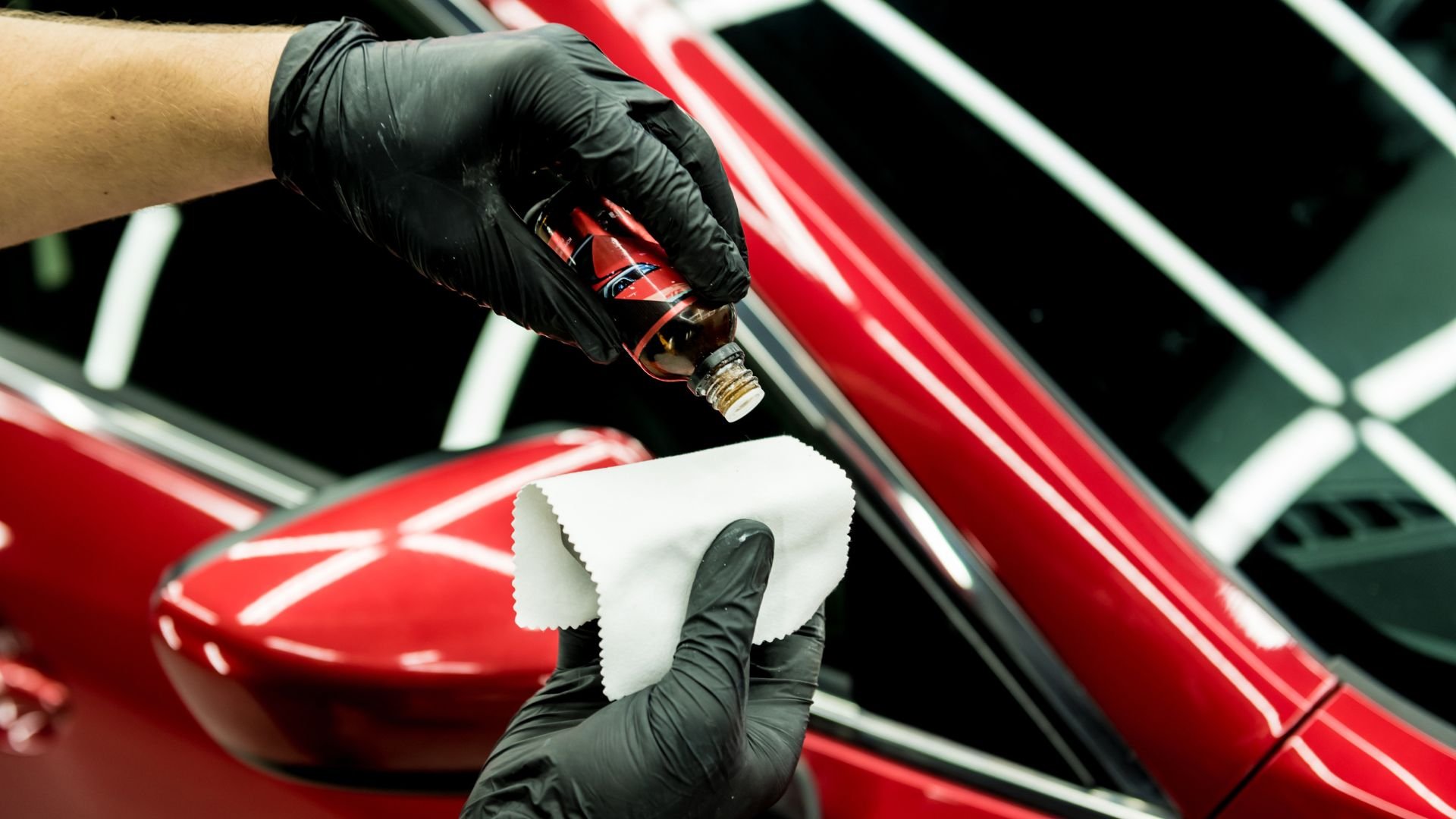 Gloved hands detailing red car with cleaning solution and cloth