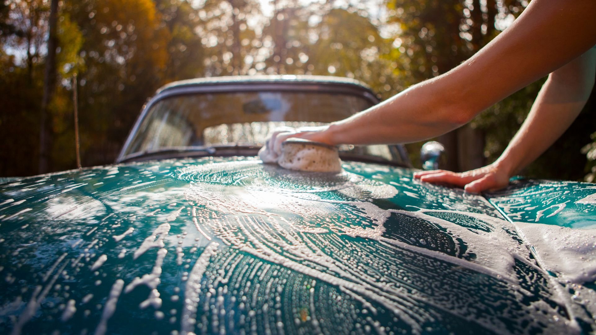 Hands washing vintage teal car with soapy sponge in autumn forest